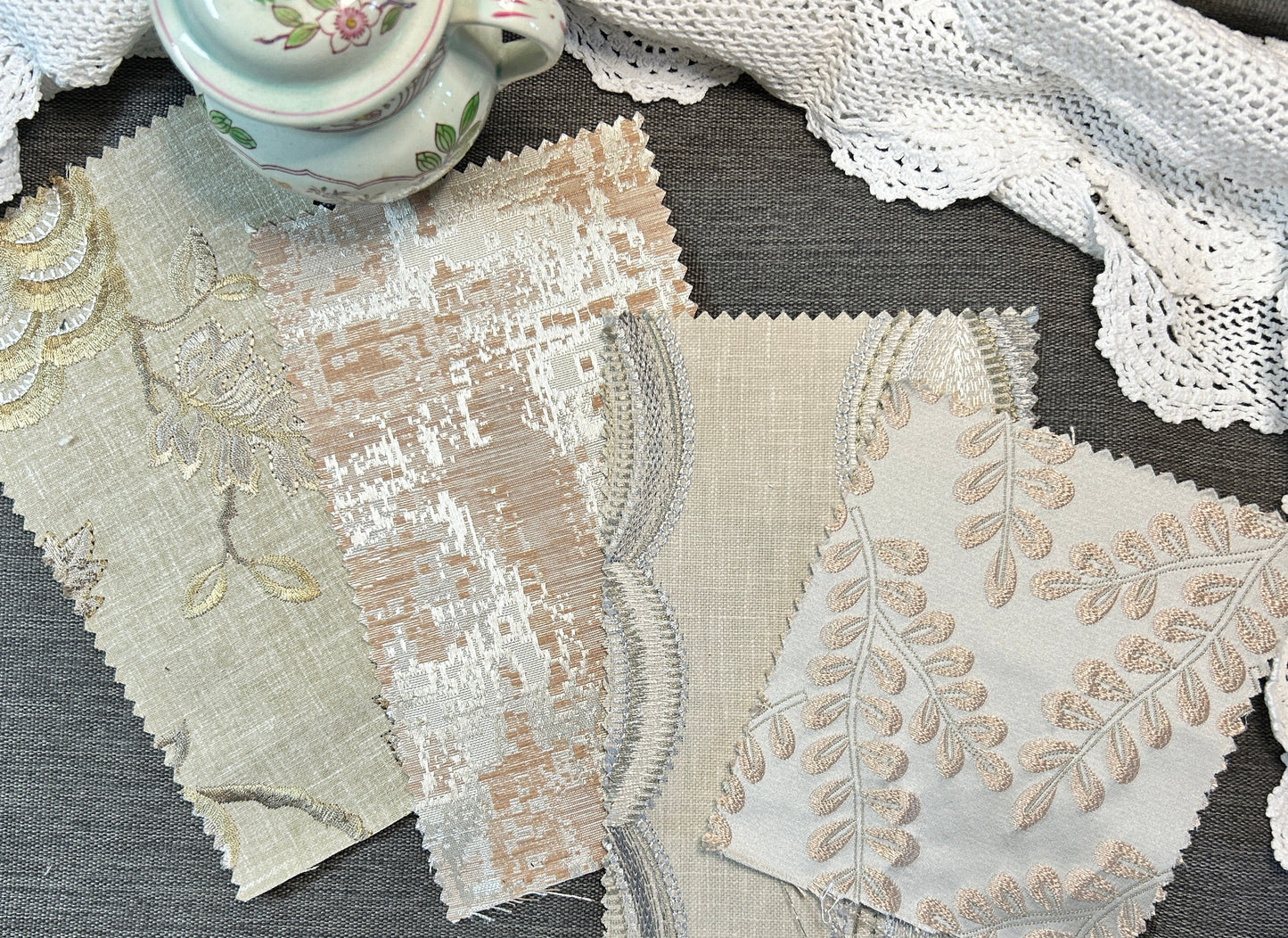 Four fabric swatches with scalloped edges and floral embroidery in beige, cream, and white tones, arranged on a gray textured surface next to a white lace doily and a small floral teacup.