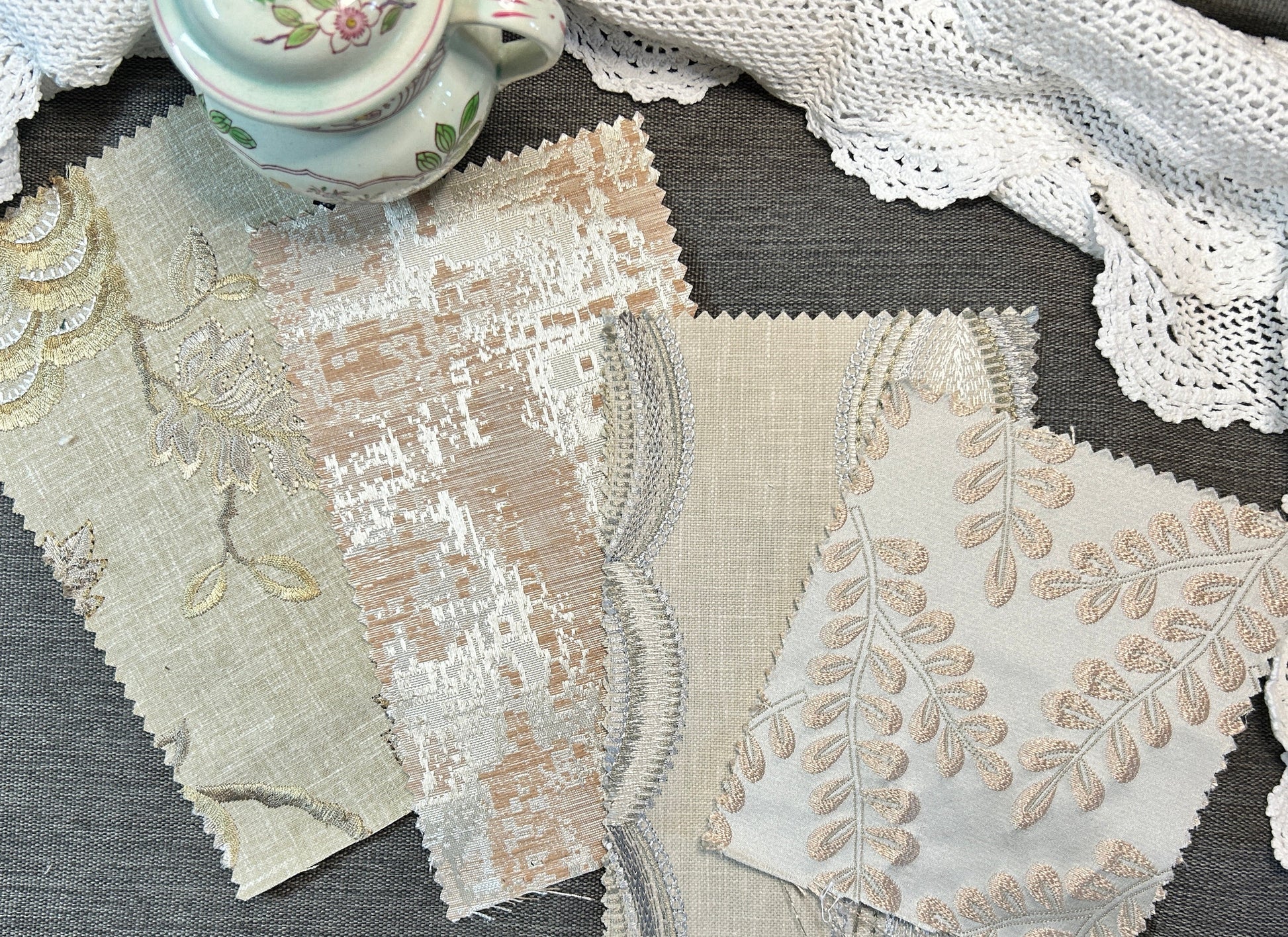 Four fabric swatches with scalloped edges and floral embroidery in beige, cream, and white tones, arranged on a gray textured surface next to a white lace doily and a small floral teacup.