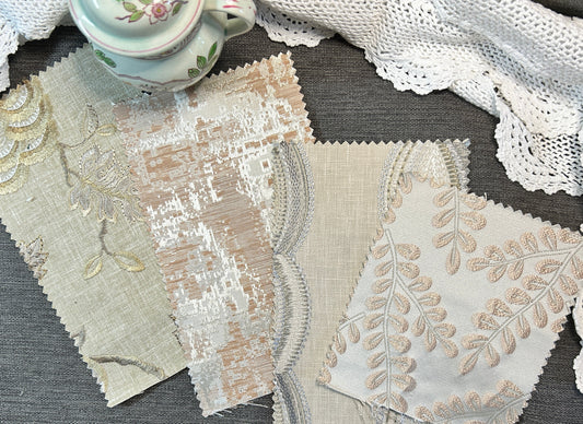 Four fabric swatches with scalloped edges and floral embroidery in beige, cream, and white tones, arranged on a gray textured surface next to a white lace doily and a small floral teacup.