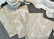 Four fabric swatches with scalloped edges and floral embroidery in beige, cream, and white tones, arranged on a gray textured surface next to a white lace doily and a small floral teacup.