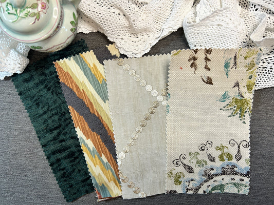 Four fabric swatches with scalloped edges laid out on a gray textured surface, surrounded by decorative white lace and a floral teacup. The swatches show different patterns: dark green textured, beige and brown ikat, light beige with silver buttons, and cream floral.