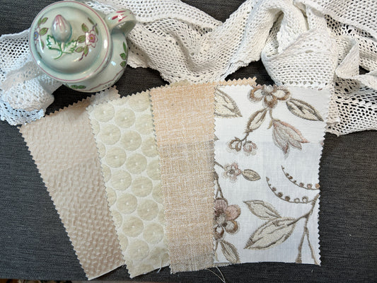 Four fabric swatches with scalloped edges laid out on a dark gray surface, alongside a white lace trim and a small floral teacup. The swatches show different textures and prints: textured cream, cream with scalloped dots, beige linen, and white with beige floral embroidery.