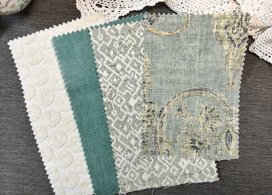 Four fabric swatches with scalloped edges laid on a dark gray textured surface. The swatches show different textile patterns and colors: white with raised circular texture, teal green, white with gray floral lace pattern, and a pale blue-gray fabric with gold embroidery.