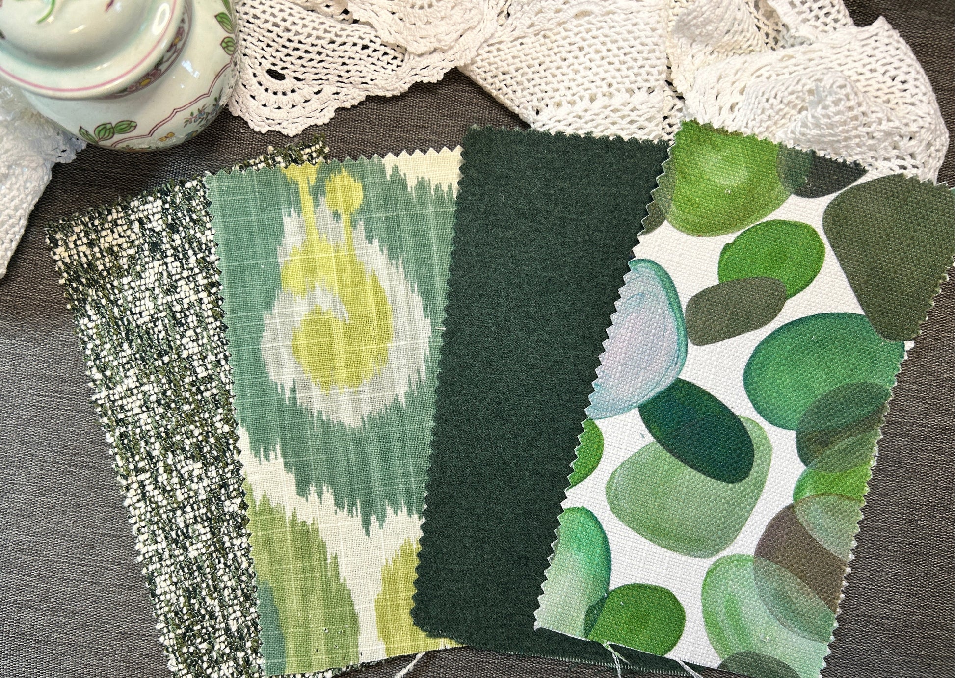Four fabric swatches with green patterns are arranged on a gray textured surface, with a lace cloth and a ceramic pot partially visible in the background.