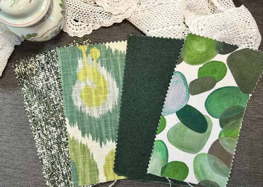 Four fabric swatches with green patterns are arranged on a gray textured surface, with a lace cloth and a ceramic pot partially visible in the background.