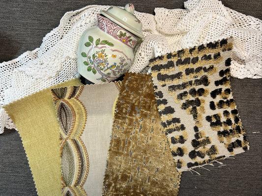 A collection of fabric and textile swatches displayed on a dark gray textured surface, alongside a white lace doily and a decorative ceramic jar. The swatches include a pale yellow and beige patterned fabric, a textured gold-brown fabric, and a beige leopard print fabric.