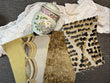 A collection of fabric and textile swatches displayed on a dark gray textured surface, alongside a white lace doily and a decorative ceramic jar. The swatches include a pale yellow and beige patterned fabric, a textured gold-brown fabric, and a beige leopard print fabric.