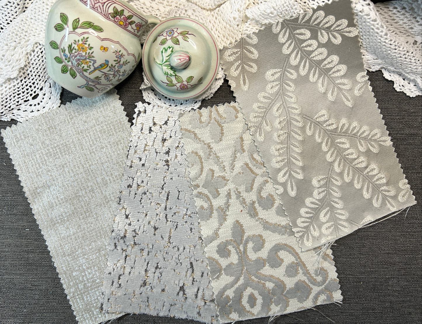 Three fabric swatches with different textures and patterns (woven, lace, and damask) in shades of gray and white, displayed on a dark gray table alongside decorative ceramic vases and white lace.