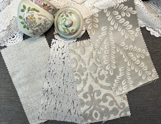 Three fabric swatches with different textures and patterns (woven, lace, and damask) in shades of gray and white, displayed on a dark gray table alongside decorative ceramic vases and white lace.