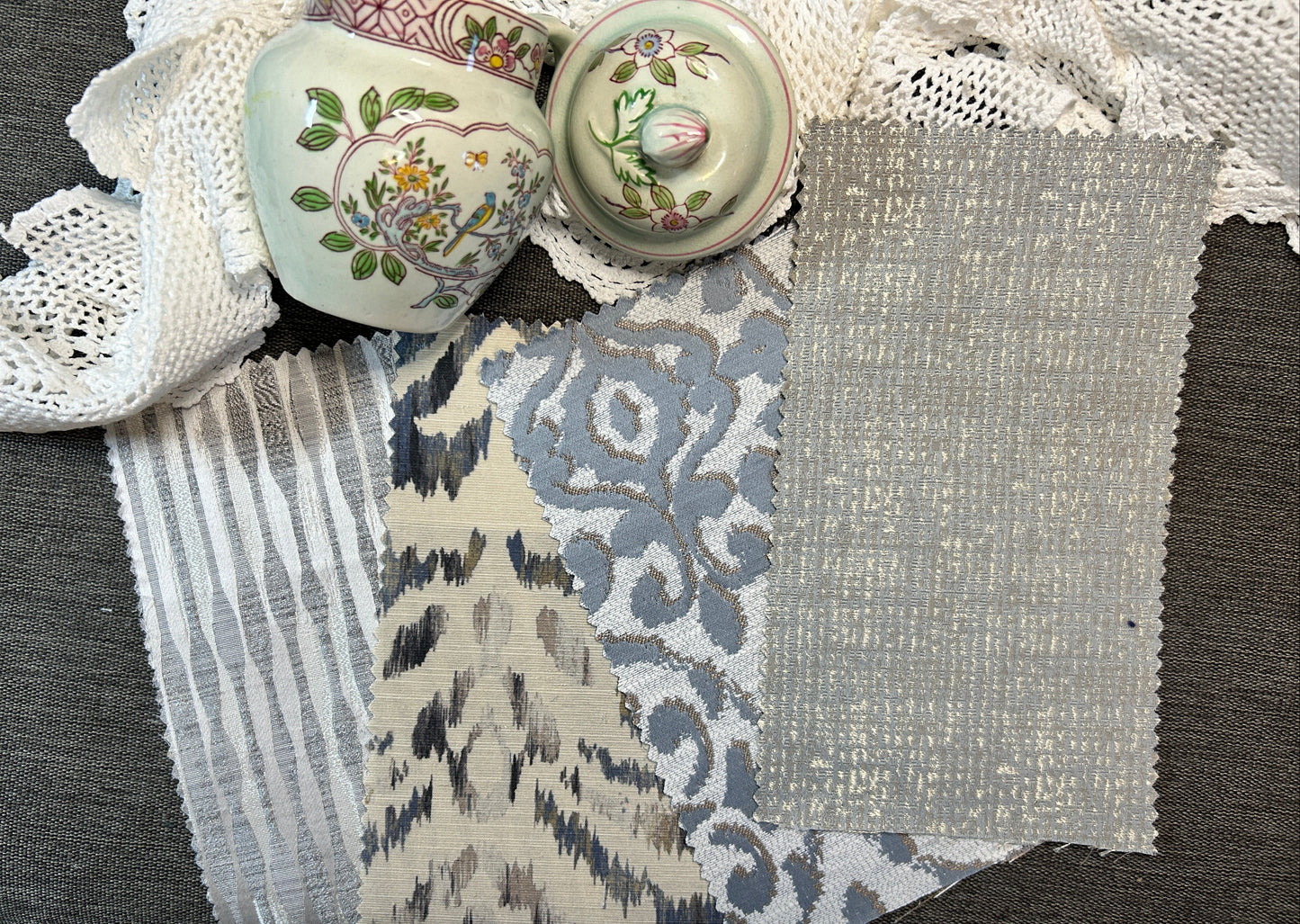 A flat-lay display of various fabric and textile swatches, including striped lace, floral ikat, and textured linen, alongside two vintage floral ceramic lidded boxes on a gray textured background.