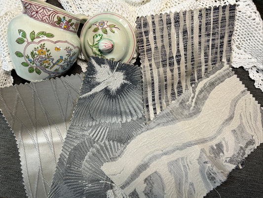 A collection of fabric and textile samples displayed on a dark gray surface with a white lace doily. The fabrics feature various patterns including gray and white abstract waves, vertical black-and-white stripes, and delicate floral embroidery on a light green ceramic pot and lid in the background.