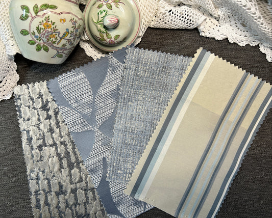 Three fabric swatches with different patterns (textured basketweave, fine horizontal stripe, and vertical stripe) in muted blue, gray, and beige tones, displayed on a dark gray textured surface next to decorative ceramic jars and a white lace cloth.